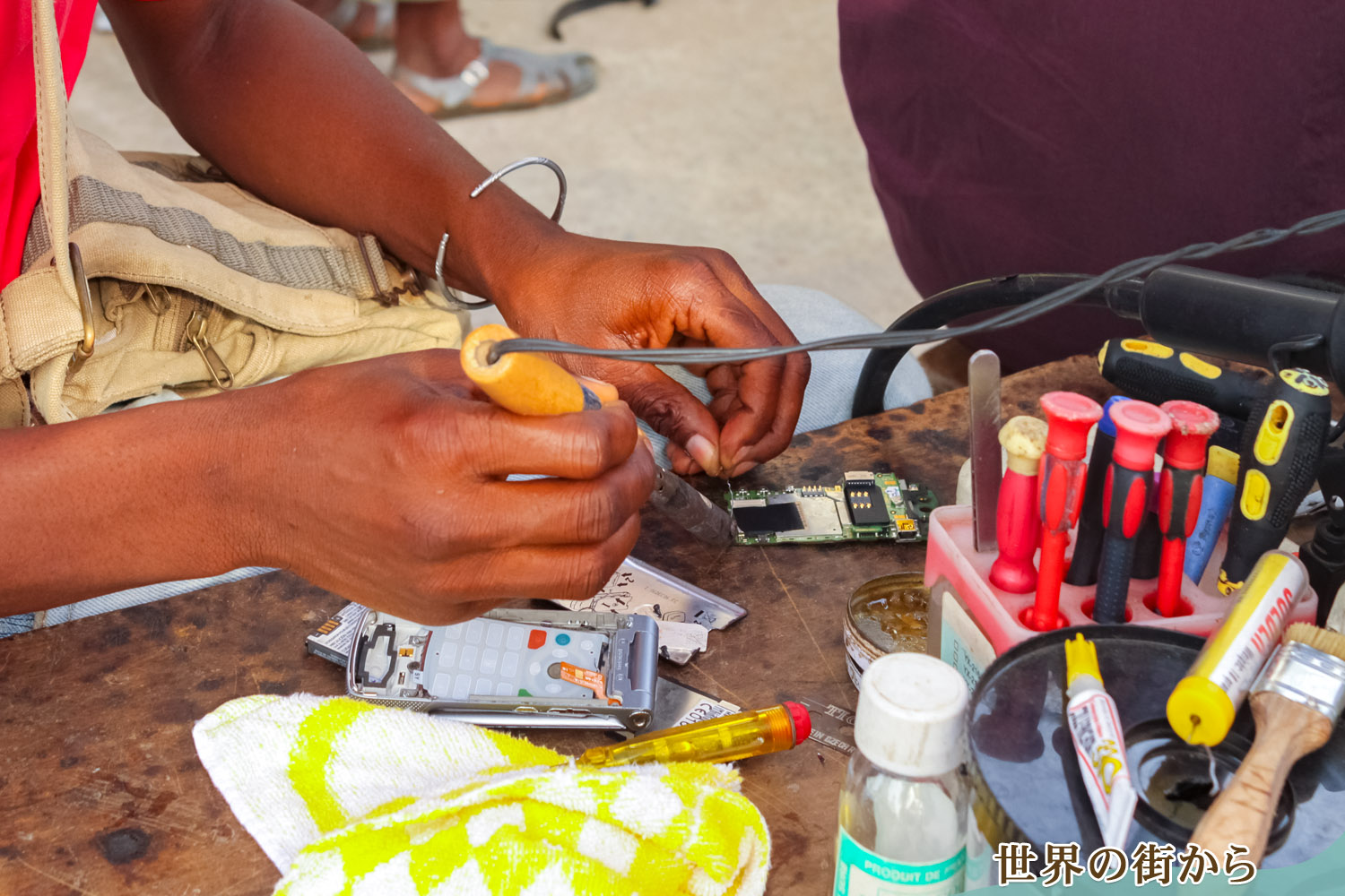 野菜や焼き魚が並ぶ市場で携帯電話を修理する熟練工